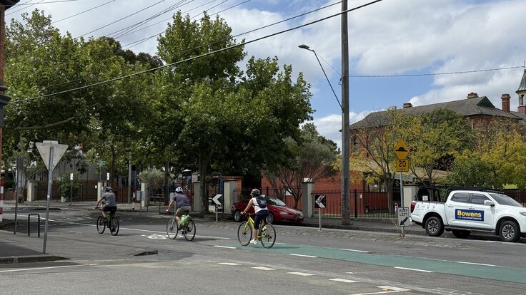 Three cyclists on the green bike lane on Wellington Street in Clifton Hill, passing the school